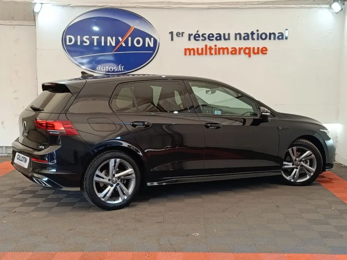 Vue de profil côté droit d'une Volkswagen Golf 1.5 eTSI noire finition R-Line avec jantes alliage, en intérieur showroom.