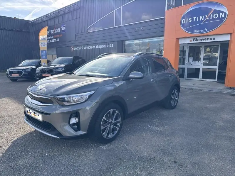 KIA Stonic brun Sienne en vue 3/4 avant droit, avec jantes alliage et barres de toit visibles, devant un bâtiment commercial.