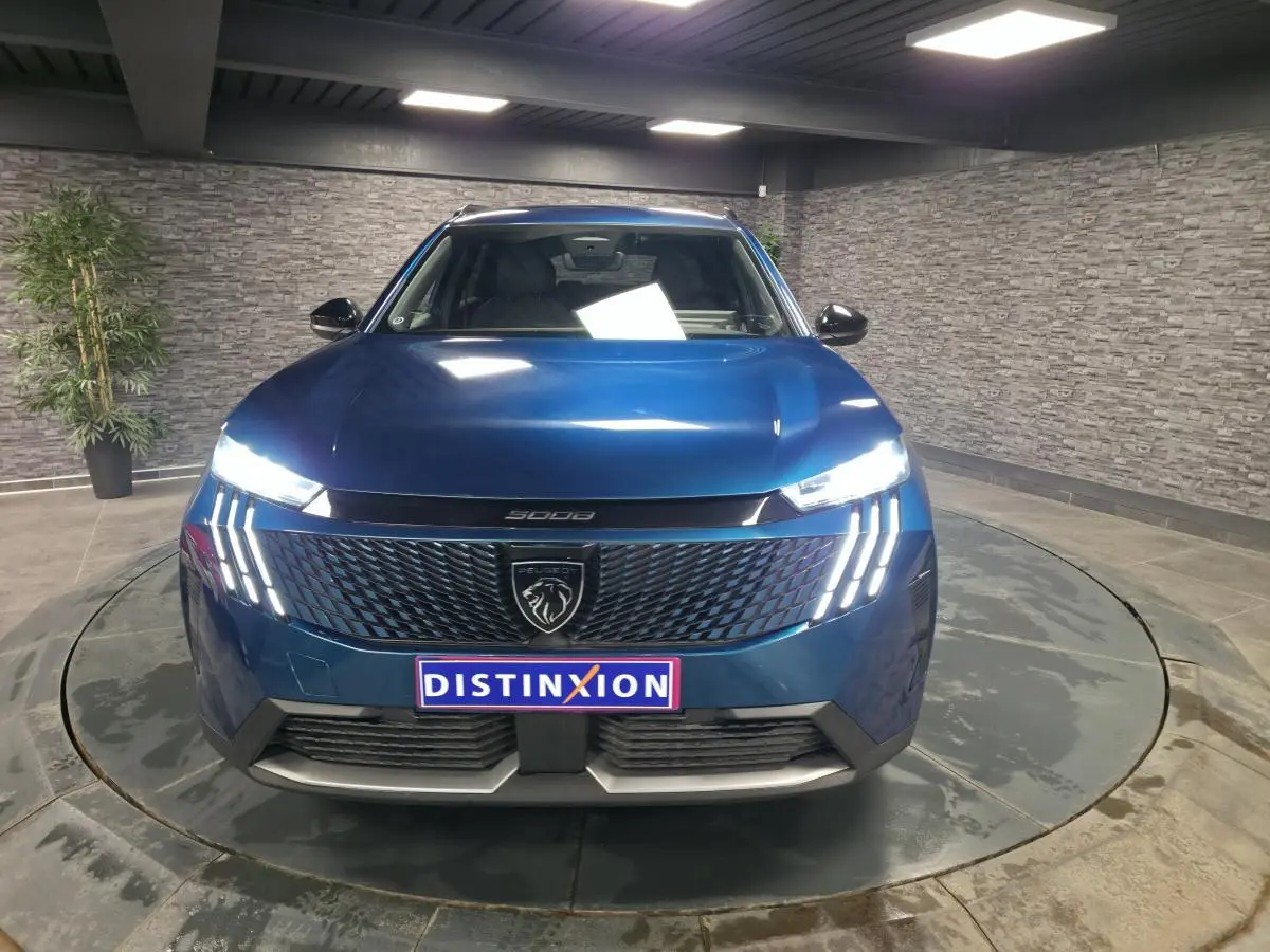 Vue avant d'un Peugeot 5008 bleu métallisé 2025 avec calandre noire et feux LED à trois griffes allumés.