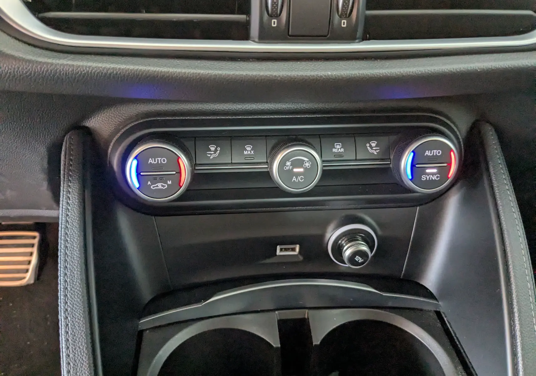 Vue rapprochée de la console centrale de l'Alfa Romeo Stelvio noir, montrant les commandes de climatisation automatique avec éclairage bleu et rouge.