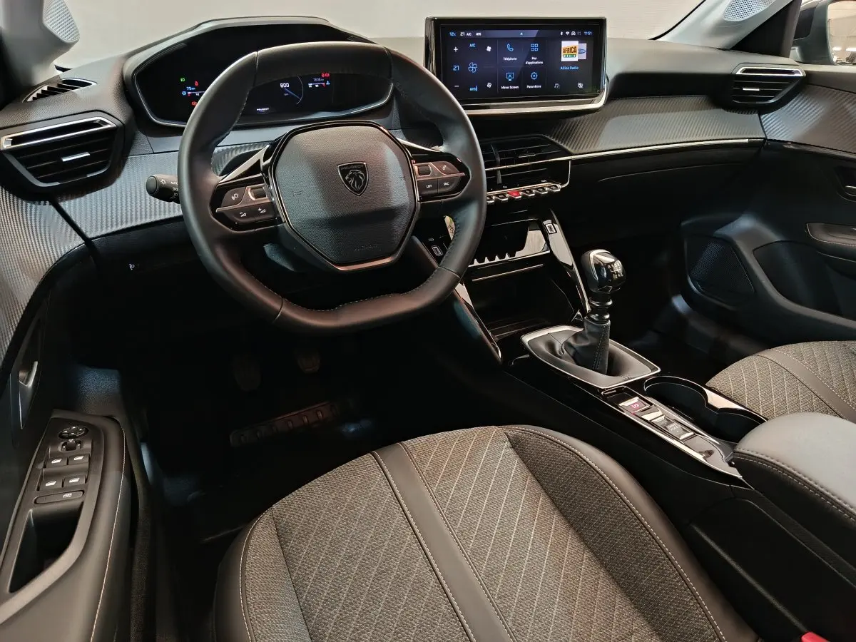 Vue intérieure avant de la Peugeot 208 gris clair, volant cuir compact, écran tactile central et boîte manuelle visible.