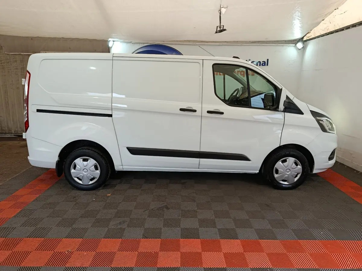 Vue de profil côté droit d’un Ford Transit Custom blanc, fourgon utilitaire avec jantes acier et protections noires sur les portières.