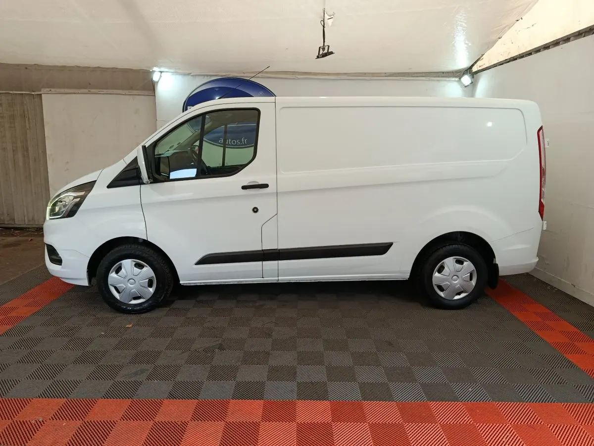 Vue de profil côté gauche d’un fourgon Ford Transit Custom blanc, avec jantes acier et habillage noir sur les bas de caisse.