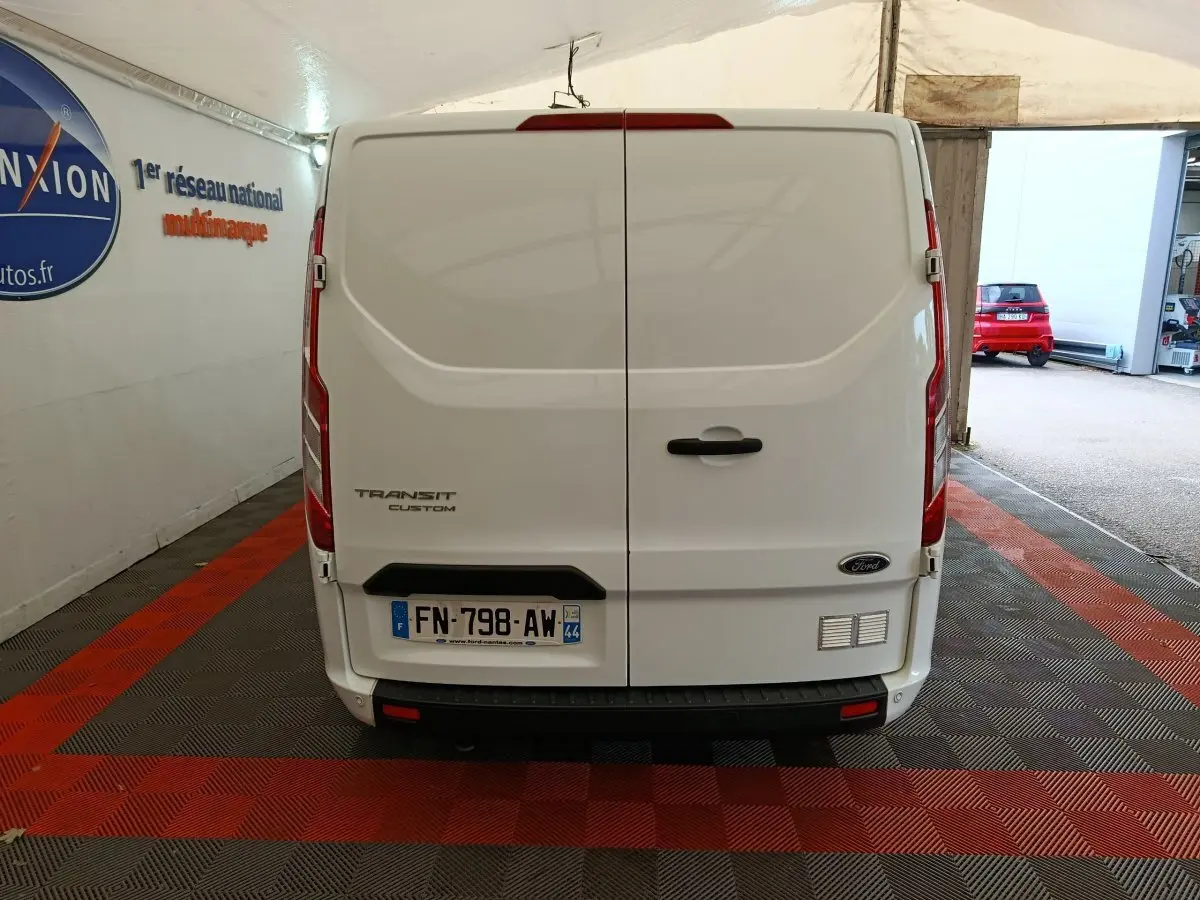 Vue arrière du fourgon blanc Ford Transit Custom, portes fermées, dans un garage avec sol à damier rouge et noir.