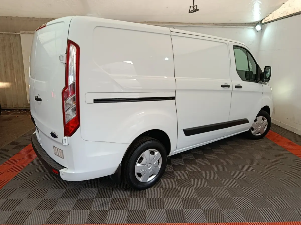 Ford Transit Custom blanc vu en 3/4 arrière droit dans un garage avec protections latérales noires et jantes acier.