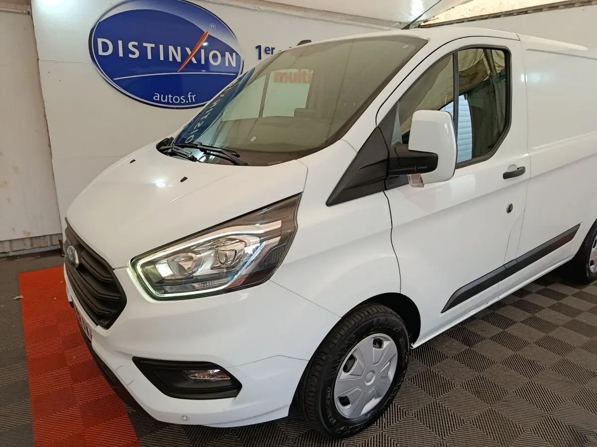 Vue 3/4 avant droit d'un fourgon Ford Transit Custom blanc avec phares allumés et jantes acier.