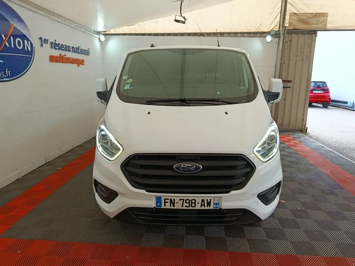 Vue avant d'un Ford Transit Custom blanc avec phares allumés dans un espace couvert.