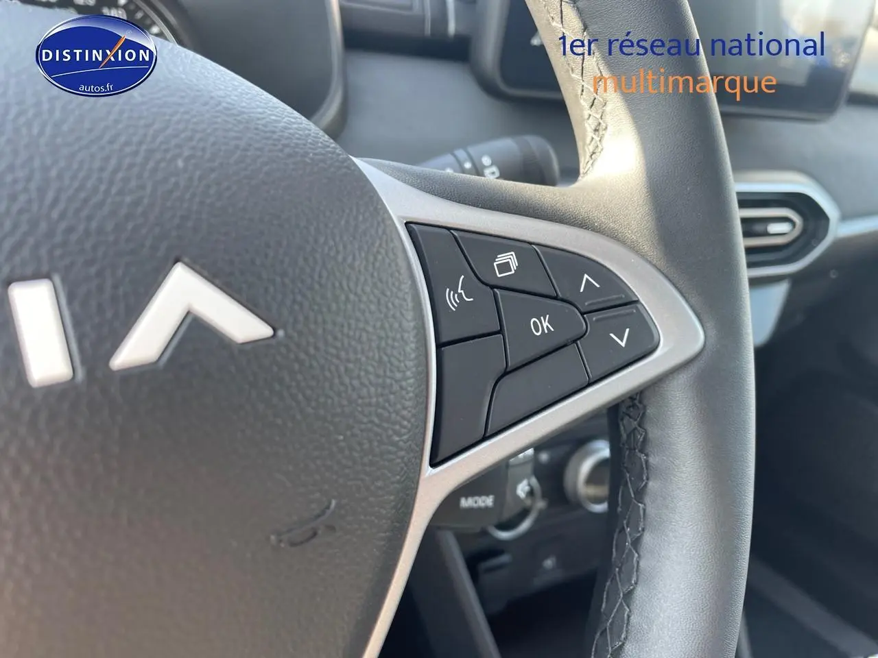 Gros plan sur les commandes au volant de la Dacia Sandero gris schiste, avec tableau de bord et aérateurs visibles en arrière-plan.
