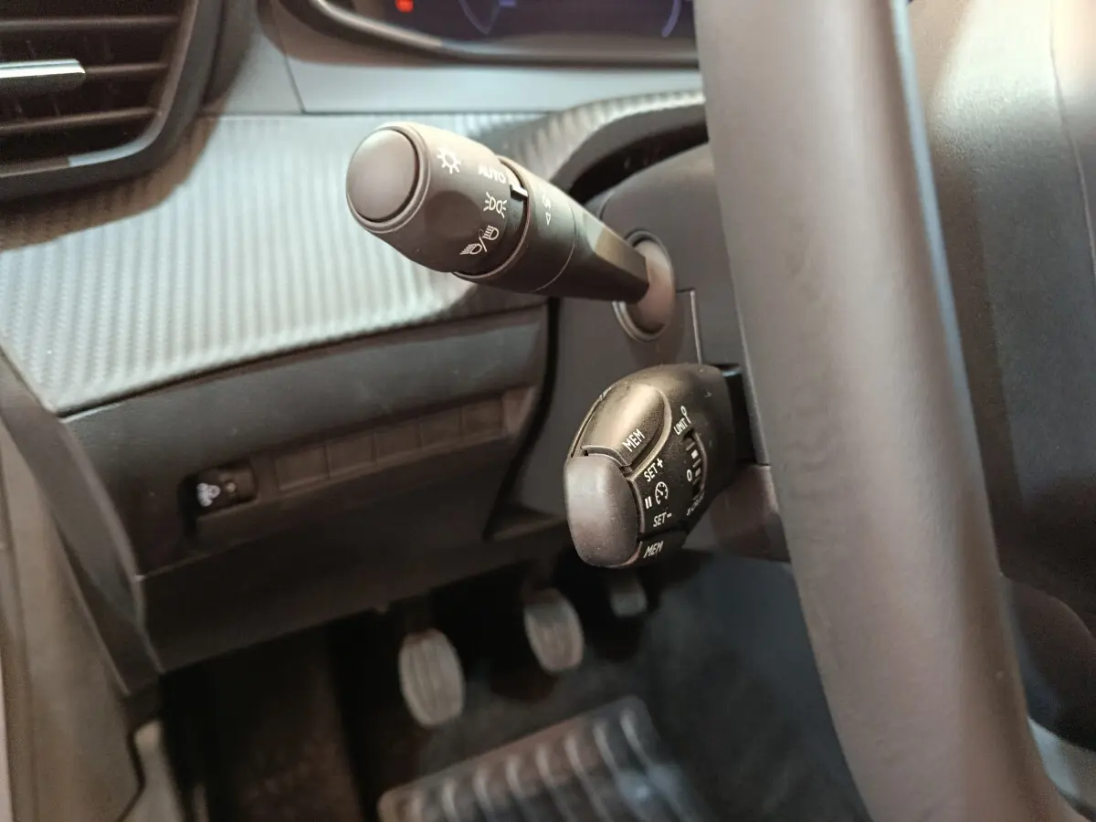 Gros plan sur les commandes au volant côté gauche du Peugeot 208 gris clair, avec volant cuir et pédales visibles.
