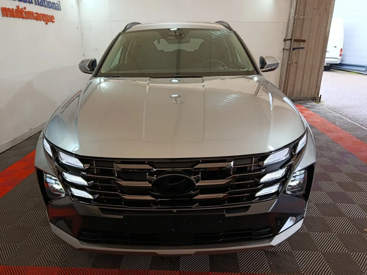 Vue avant d’un Hyundai Tucson gris clair 2025 avec calandre large et phares LED intégrés, en intérieur showroom.