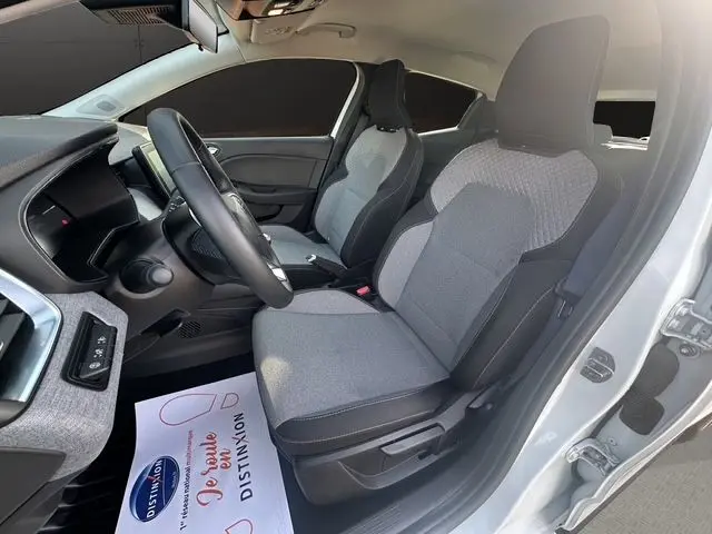 Vue intérieure côté conducteur d'une Renault Clio V blanche, sièges tissu gris à motif 3D et volant textile grainé.