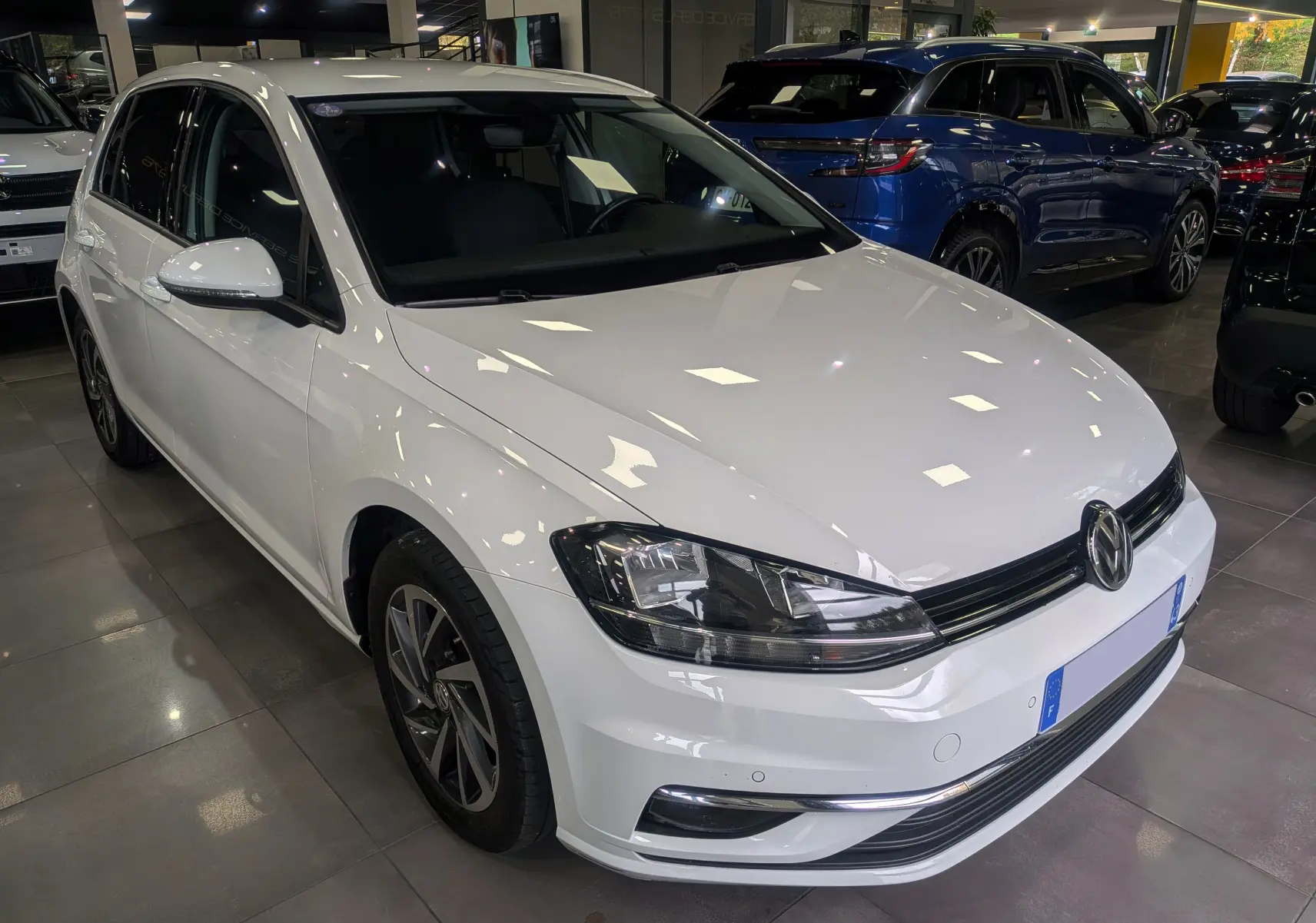 Volkswagen Golf VII TSI blanche vue en 3/4 avant droit dans un showroom avec jantes alliage et phares LED.