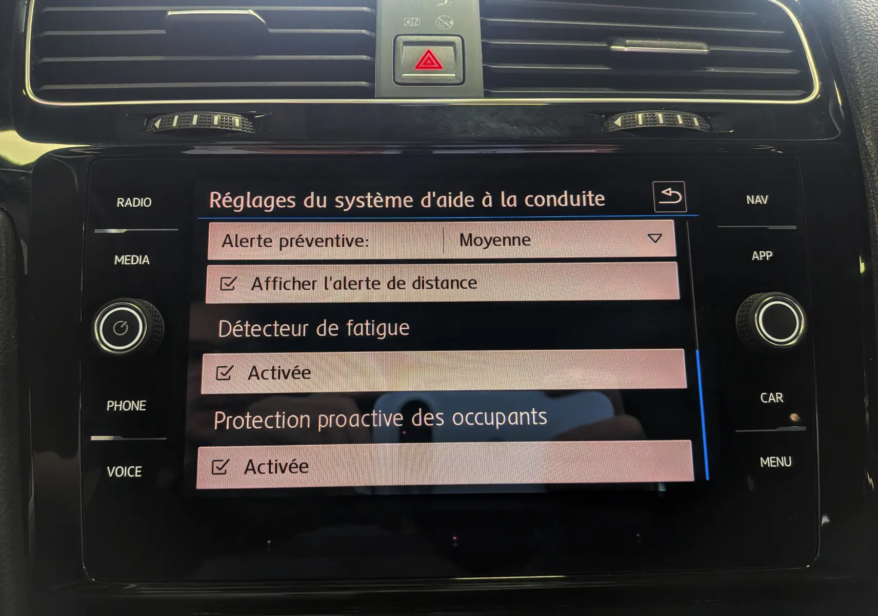 Vue rapprochée de l'écran tactile central noir du Volkswagen Golf VII blanc, affichant les réglages d'aide à la conduite.