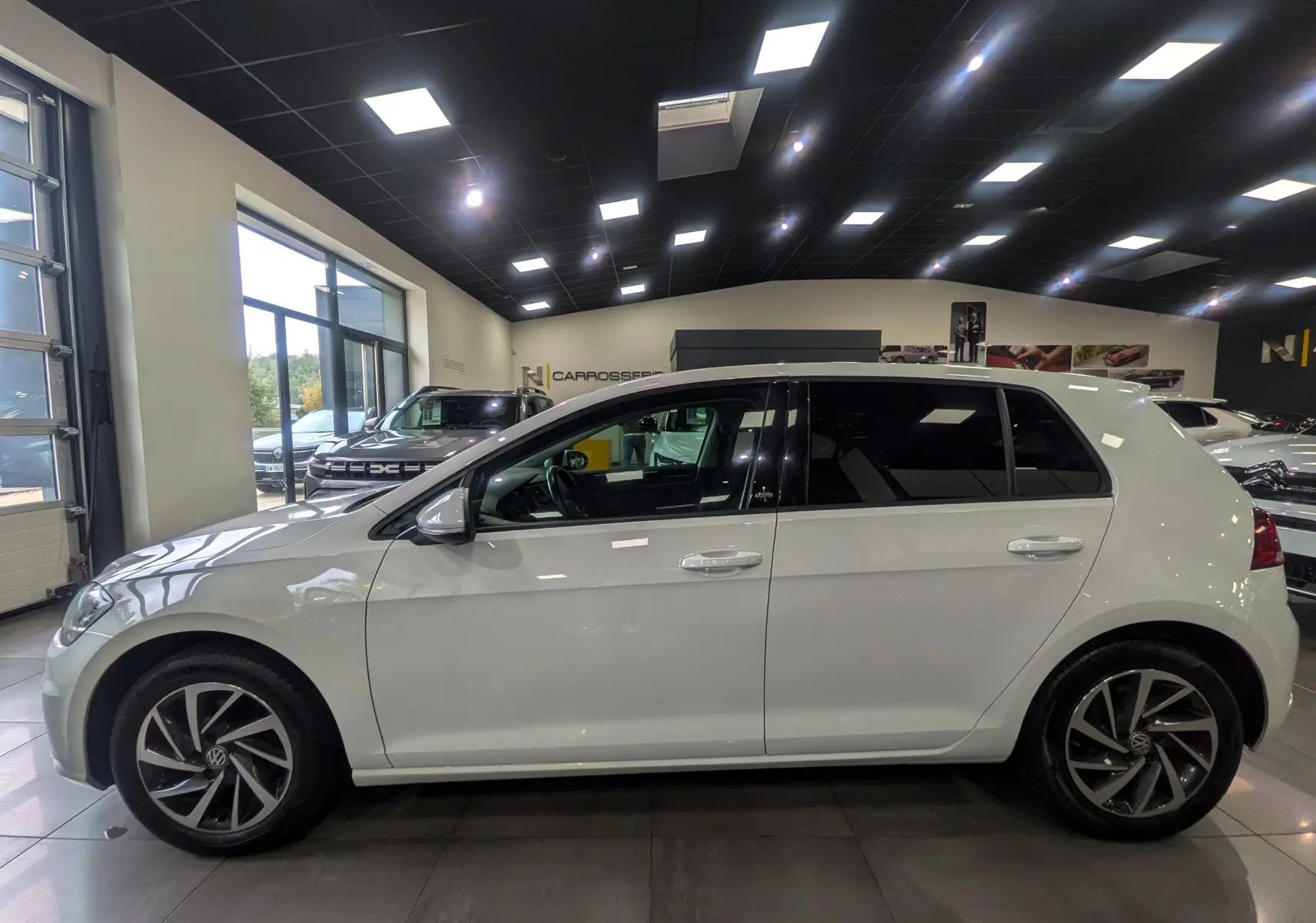 Profil côté gauche d'une Volkswagen Golf VII TSI 125 CH Sound blanche, avec jantes alliage et vitres teintées en showroom.