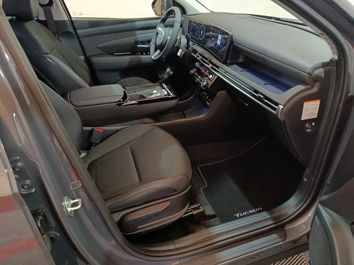 Vue intérieure côté conducteur du Hyundai Tucson gris foncé 2025, avec tableau de bord numérique et sièges tissu noirs.