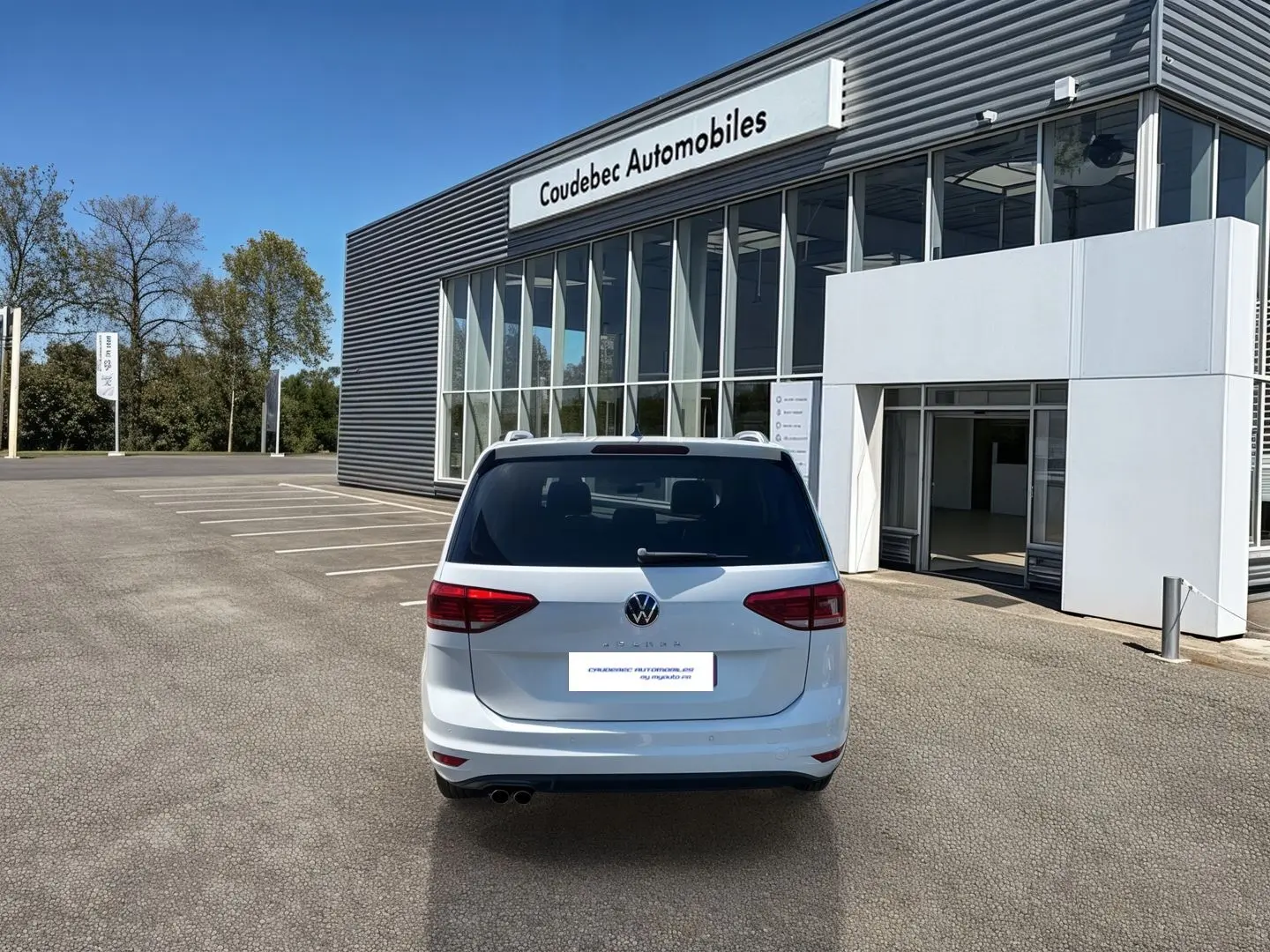 Vue arrière d'un Volkswagen Touran blanc 2025 stationné devant un bâtiment de concession Coudebec Automobiles.