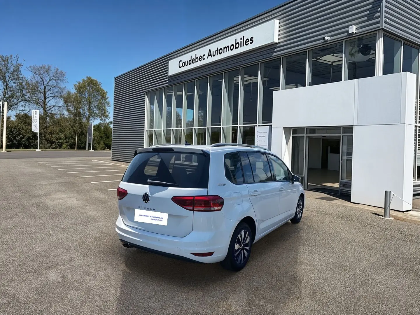 Volkswagen Touran blanc vu en 3/4 arrière droit devant un bâtiment de concession Coudebec Automobiles.