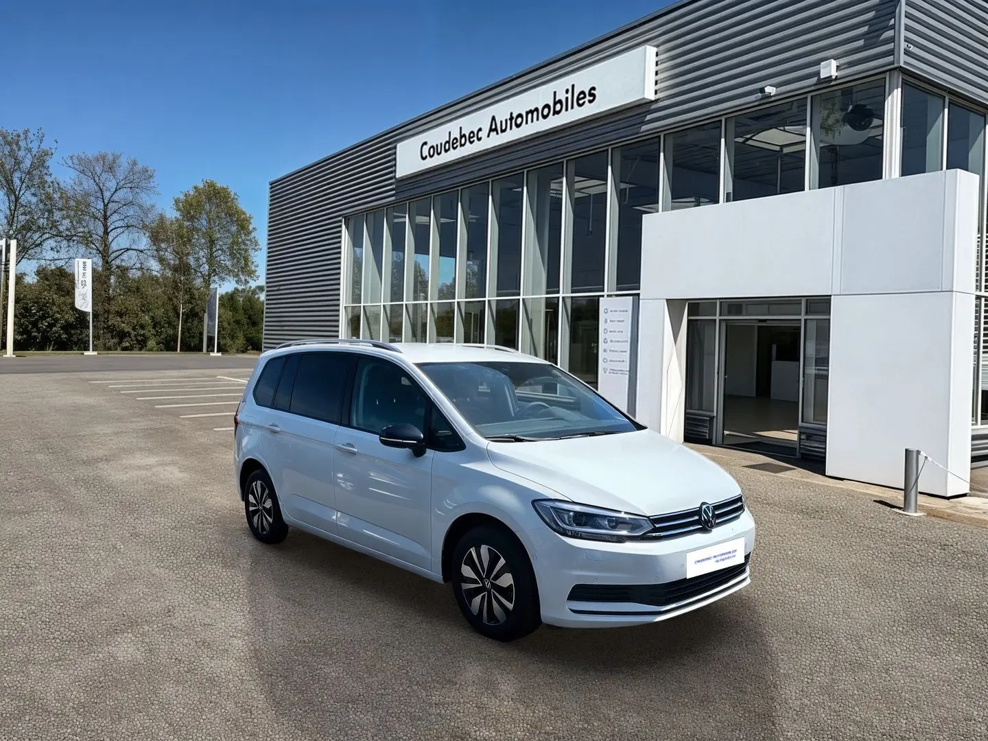 Volkswagen Touran blanc vu en 3/4 avant droit devant un bâtiment moderne de concession automobile.