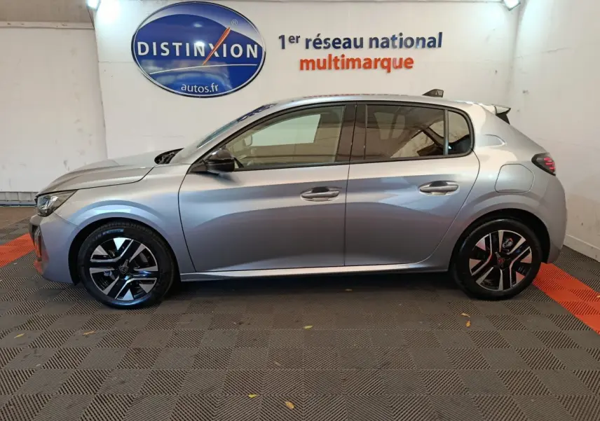 Profil côté gauche d'une Peugeot 208 gris clair 2025 garée en intérieur avec jantes alliage noires et logo Distinxion en arrière-plan.