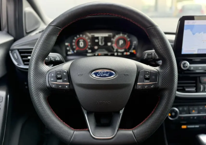 Gros plan sur le volant ST-Line en cuir perforé avec surpiqûres rouges du Ford Puma 2024, tableau de bord numérique et écran GPS visibles.