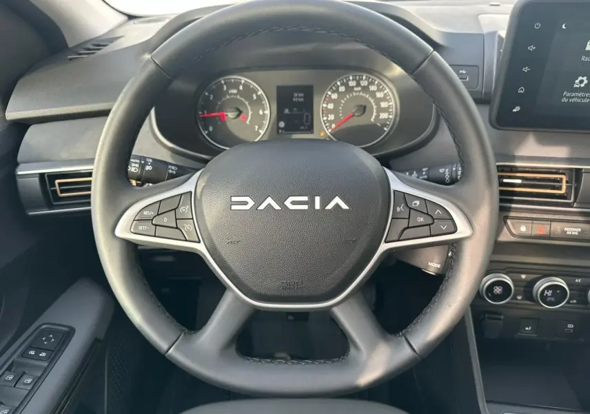 Vue rapprochée du volant noir Dacia avec commandes intégrées et tableau de bord numérique en arrière-plan.