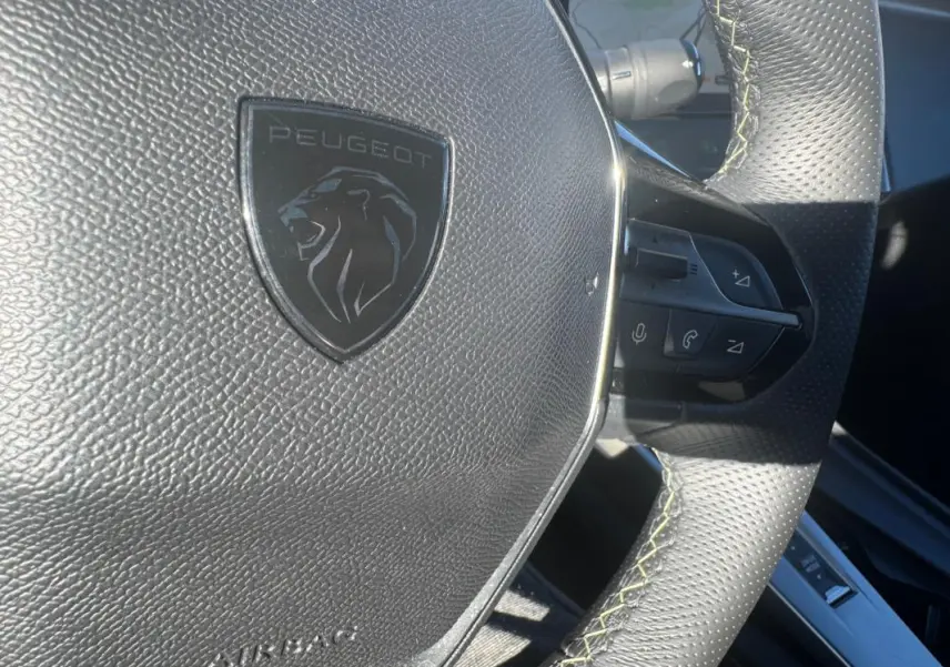 Gros plan sur le volant cuir noir perforé de la Peugeot 408 Hybrid 136 e-DCS6 GT avec logo lion et commandes intégrées.