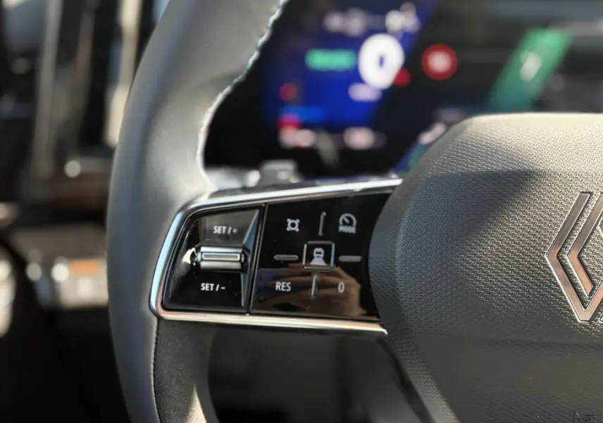 Gros plan sur les commandes gauche du volant gris texturé du Renault Austral E-Tech, avec tableau de bord numérique flou en arrière-plan.
