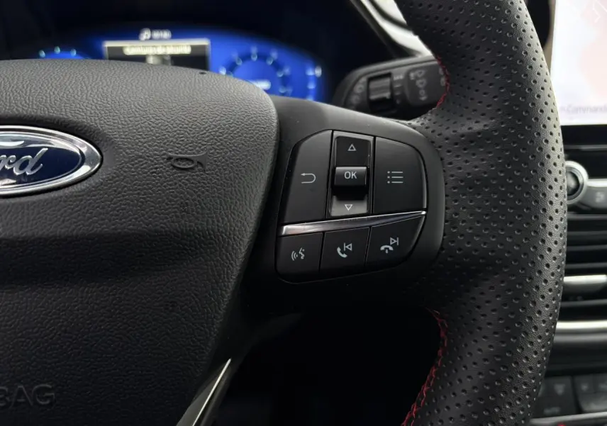 Gros plan sur le volant perforé noir avec surpiqûres rouges du Ford Puma 2024, boutons multifonctions visibles.