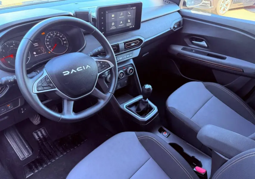 Vue intérieure côté conducteur de la Dacia Sandero 2025, volant multifonctions, écran tactile et sièges tissu brun cuivré.