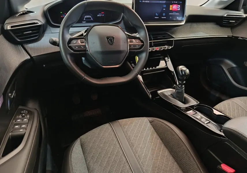 Intérieur de la Peugeot 208 PureTech 100 S&S Allure 2025, vue du poste de conduite avec volant cuir et écran tactile central.