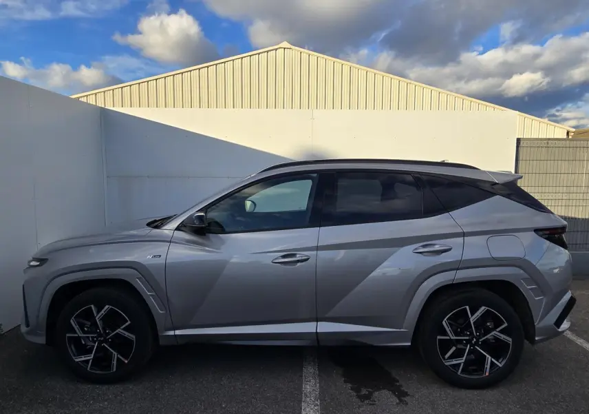 Vue de profil côté gauche d'un Hyundai Tucson 2025 gris métallisé avec jantes alliage noires et toit noir.