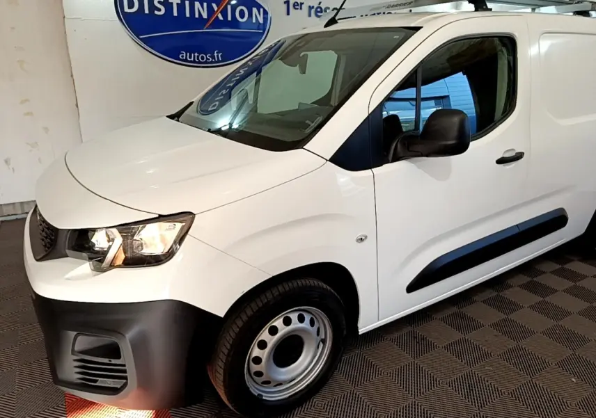 Vue 3/4 avant droit d’un Peugeot Partner blanc avec pare-chocs noir et barres de toit en intérieur showroom.