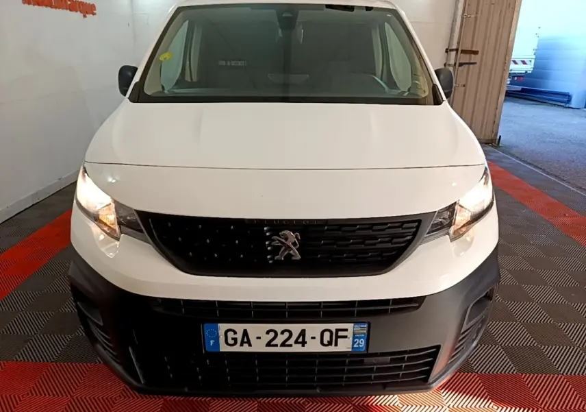 Vue avant d’un Peugeot Partner blanc 2021 avec calandre noire et phares allumés dans un garage.