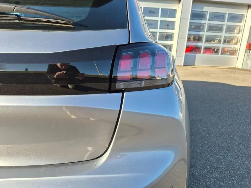 Gros plan sur le feu arrière droit d'une Peugeot 208 gris Artense, avec reflet du photographe sur la carrosserie.