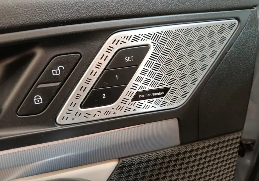 Détail intérieur côté conducteur avec commandes de verrouillage et haut-parleur Harman Kardon argenté sur porte noire.
