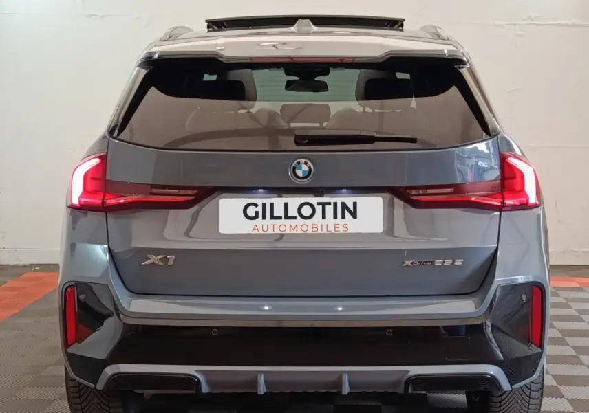 Vue arrière d’un BMW X1 xDrive25e M Sport gris foncé avec feux LED et logo visible sur fond neutre intérieur.