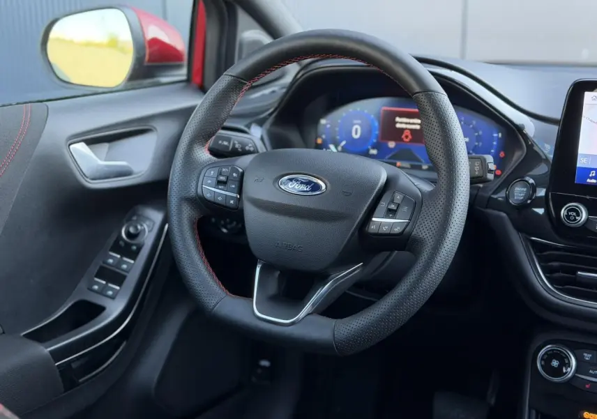 Vue intérieure centrée sur le volant ST-Line noir avec surpiqûres rouges, tableau de bord numérique et commandes sur porte côté gauche.