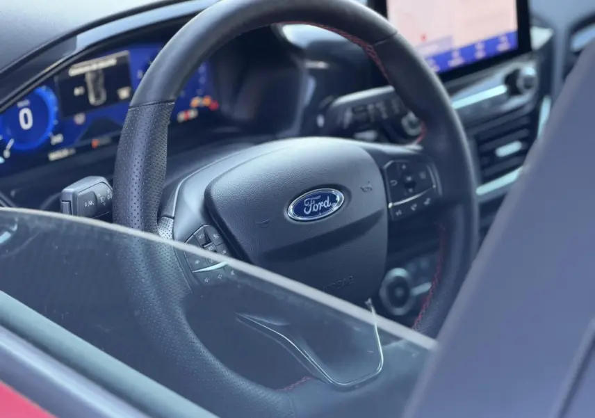Vue rapprochée de l’intérieur du Ford Puma 2023, volant ST-Line en cuir noir avec surpiqûres rouges visible à travers la vitre partiellement baissée.
