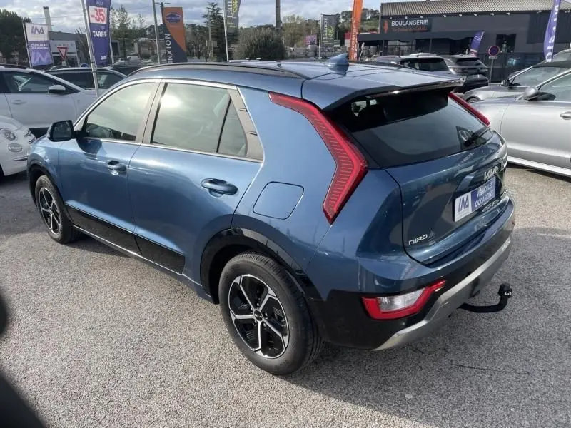 Vue 3/4 arrière droite du Kia Niro 2024 bleu métal, hybride essence, avec feux arrière en forme de C et attelage visible.