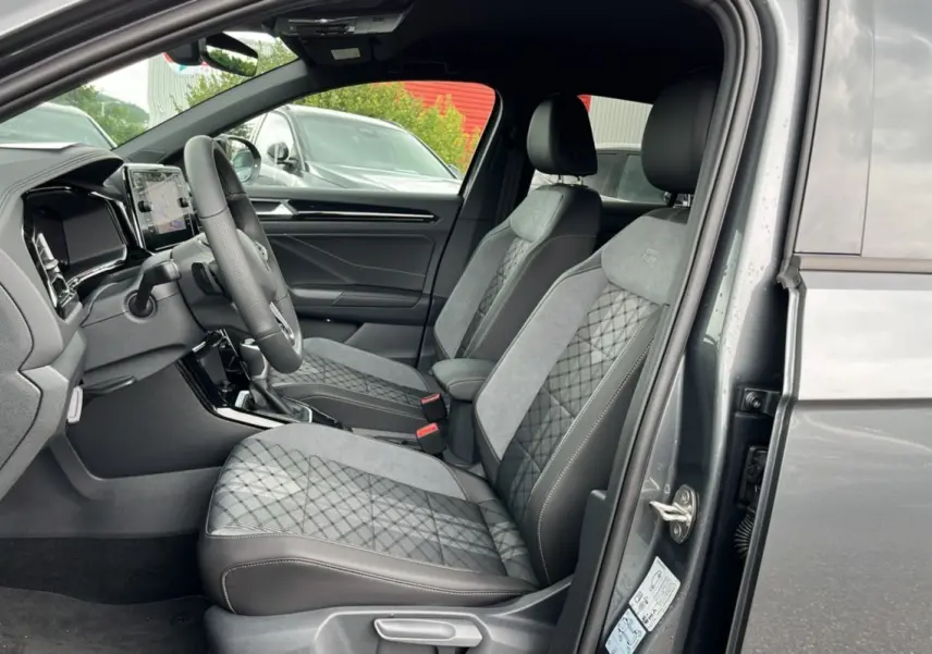 Vue intérieure côté conducteur du Volkswagen T-Roc gris indium 2025, sièges tissu R-Line avec surpiqûres et volant cuir multifonctions.
