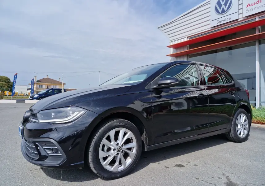 Volkswagen Polo noire vue en 3/4 avant droit, garée devant un service Volkswagen avec jantes argentées.