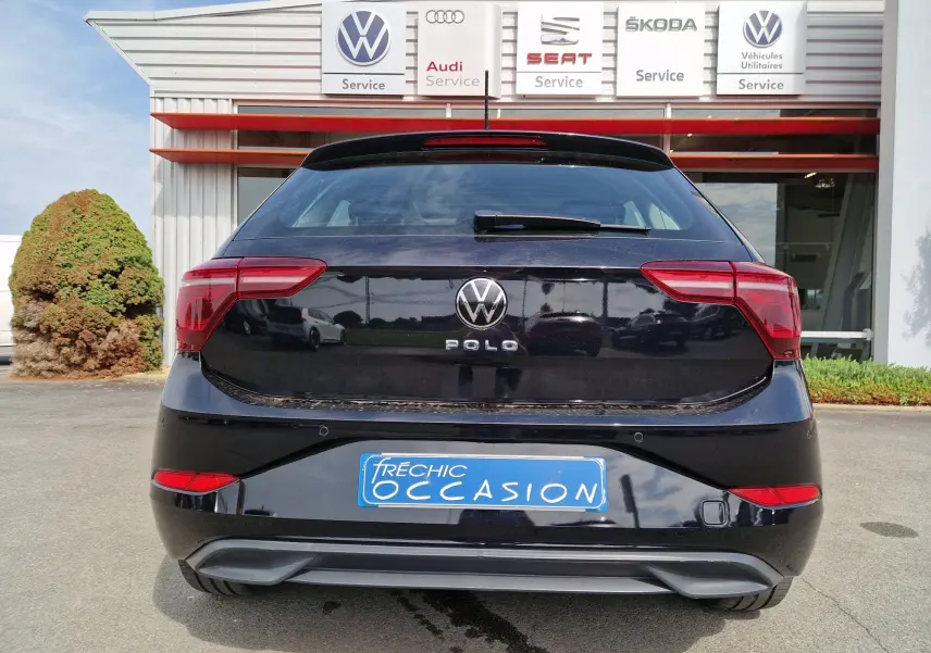 Vue arrière d'une Volkswagen Polo noire 2022 stationnée devant un garage multimarques avec plaque d'occasion bleue.