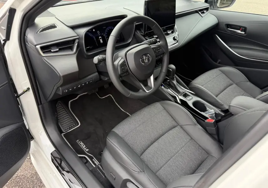 Vue intérieure côté conducteur de la Toyota Corolla Touring Sport blanche, avec tableau de bord moderne et sièges gris tissu et cuir.