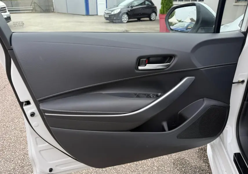 Porte avant gauche ouverte d'une Toyota Corolla Touring Sport blanche, intérieur noir avec poignée argentée et commandes de vitres.