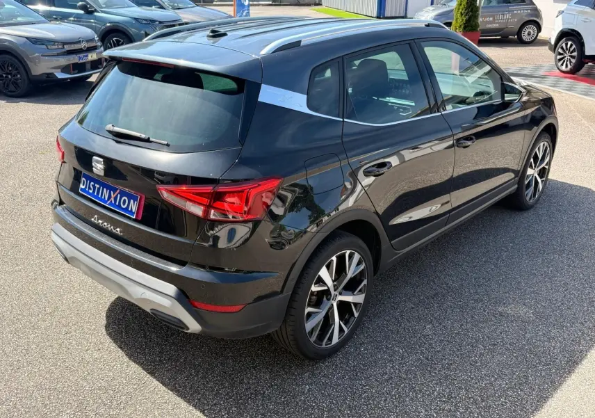 Vue 3/4 arrière droite d'un SEAT Arona noir avec jantes alliage et barres de toit chromées sur parking.