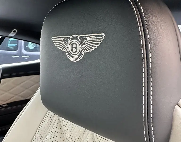 Gros plan sur l'appui-tête noir brodé du logo Bentley avec surpiqûres blanches, siège cuir beige perforé intérieur Continental GT.