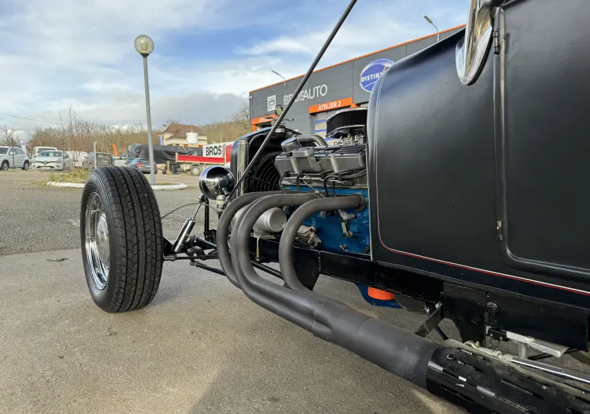 Vue de profil avant droit du Ford T Hot Rod noir, mettant en valeur son moteur V8 bleu et ses échappements latéraux noirs.