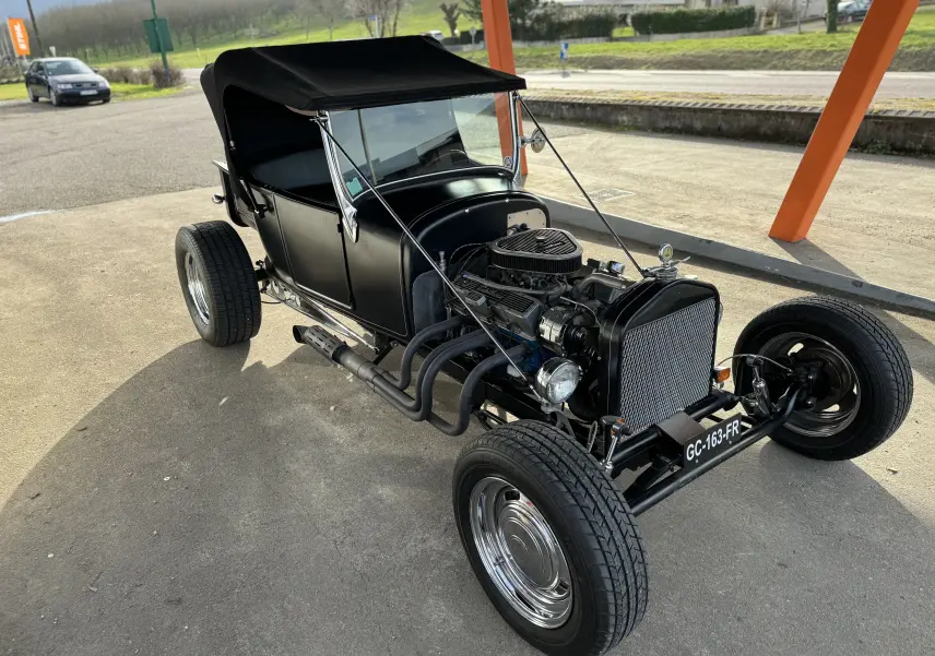 Ford T Hot Rod V8 noir vue 3/4 avant, capote relevée et moteur V8 apparent avec échappements latéraux.