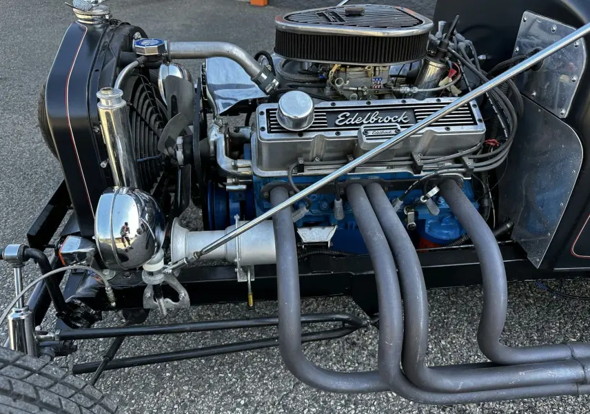Gros plan sur le moteur V8 Edelbrock bleu et gris du Ford T Hot Rod noir, avec échappements latéraux visibles.