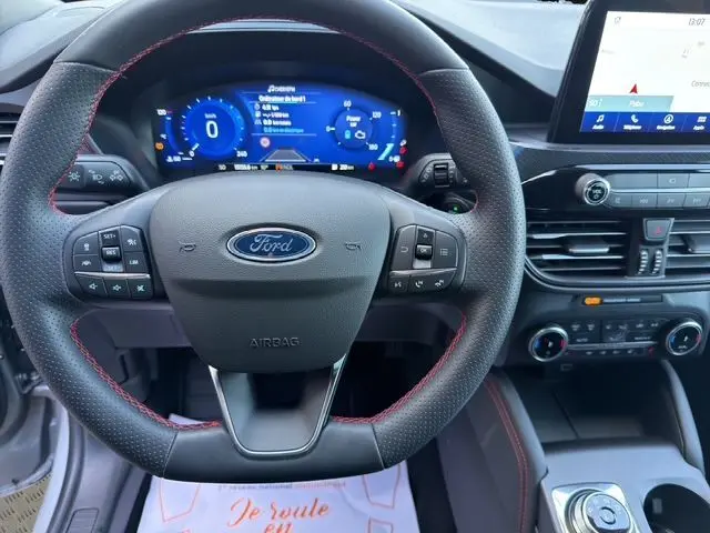 Vue intérieure centrée sur le volant noir avec surpiqûres rouges du Ford Kuga 2024 hybride, tableau de bord digital et écran tactile.
